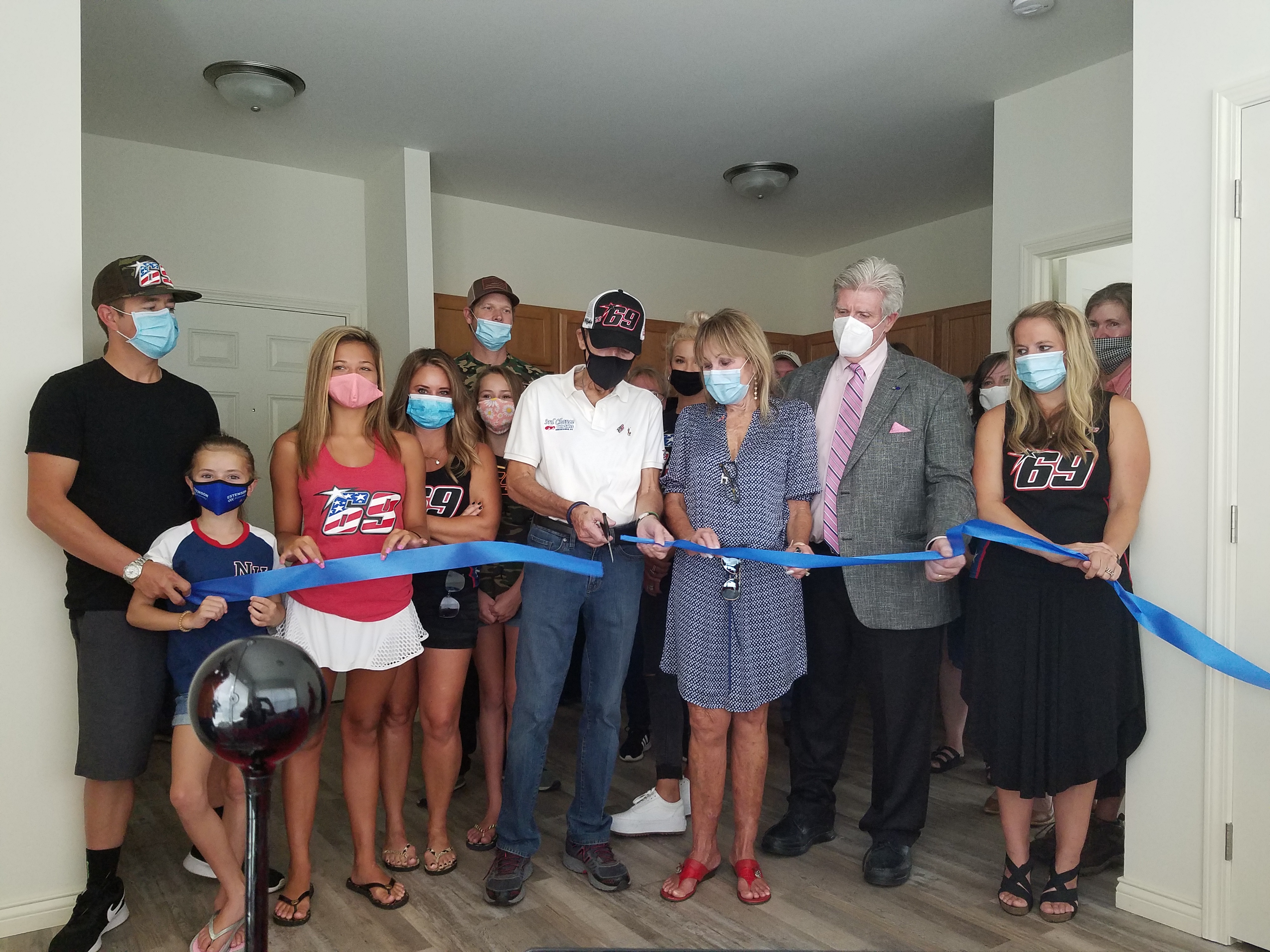 A group of people, most wearing masks, stand indoors as two individuals cut a blue ribbon with large scissors during a ribbon-cutting ceremony.