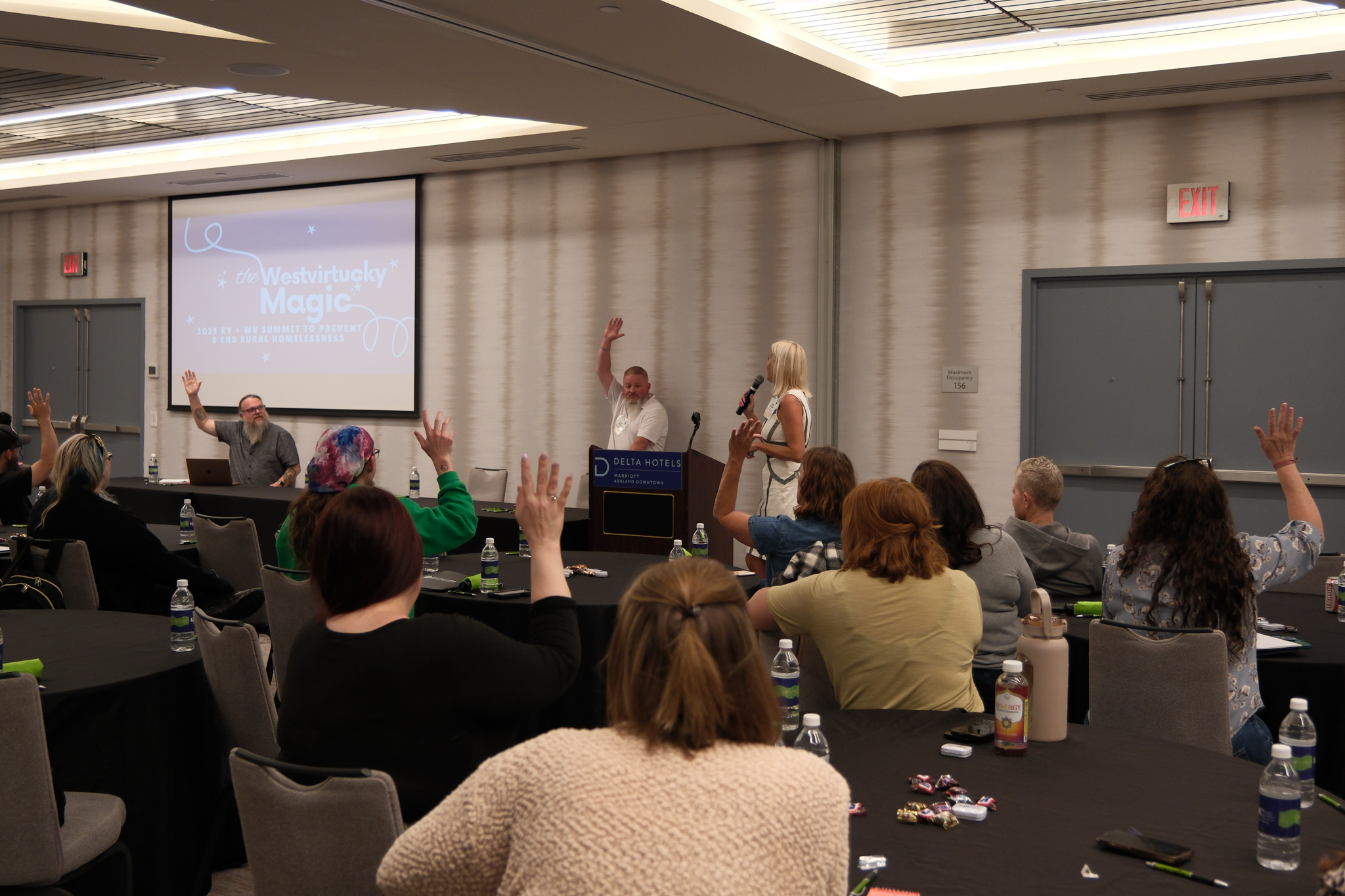 A group of people seated at tables in a conference room raise their hands while a presenter speaks at a podium. A projection screen displays “Westirucky Magic.”.