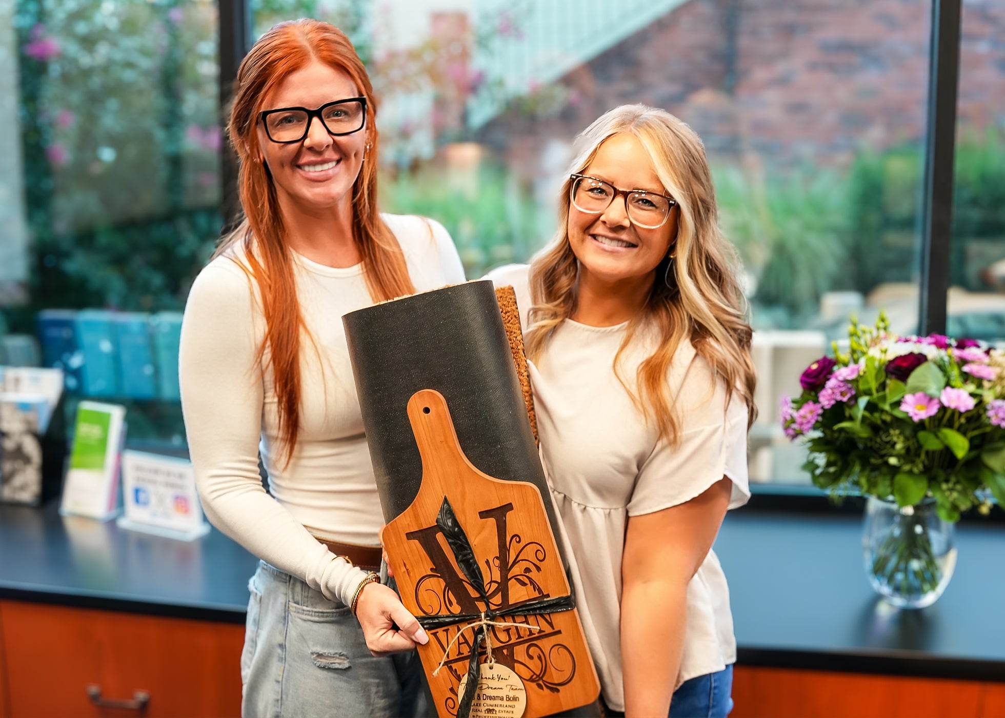 Amanda with her real estate agent, Dreama Bolin. The two women stand indoors, smiling at the camera. One holds a wooden award shaped like a cutting board. A vase of purple flowers is on the counter beside them, with windows in the background.