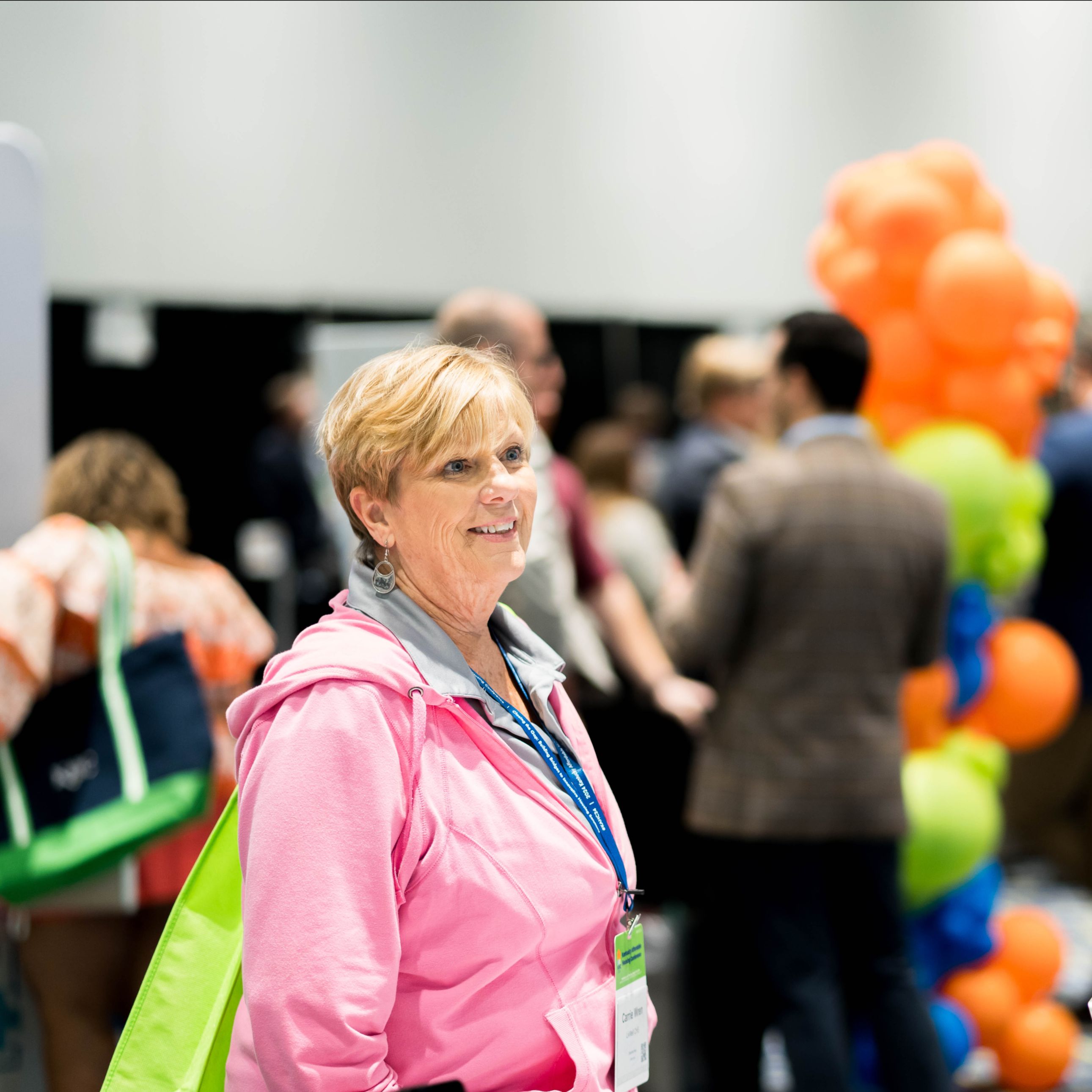 A woman in a pink jacket with a green tote bag stands indoors at a crowded event with colorful balloon decorations in the background.