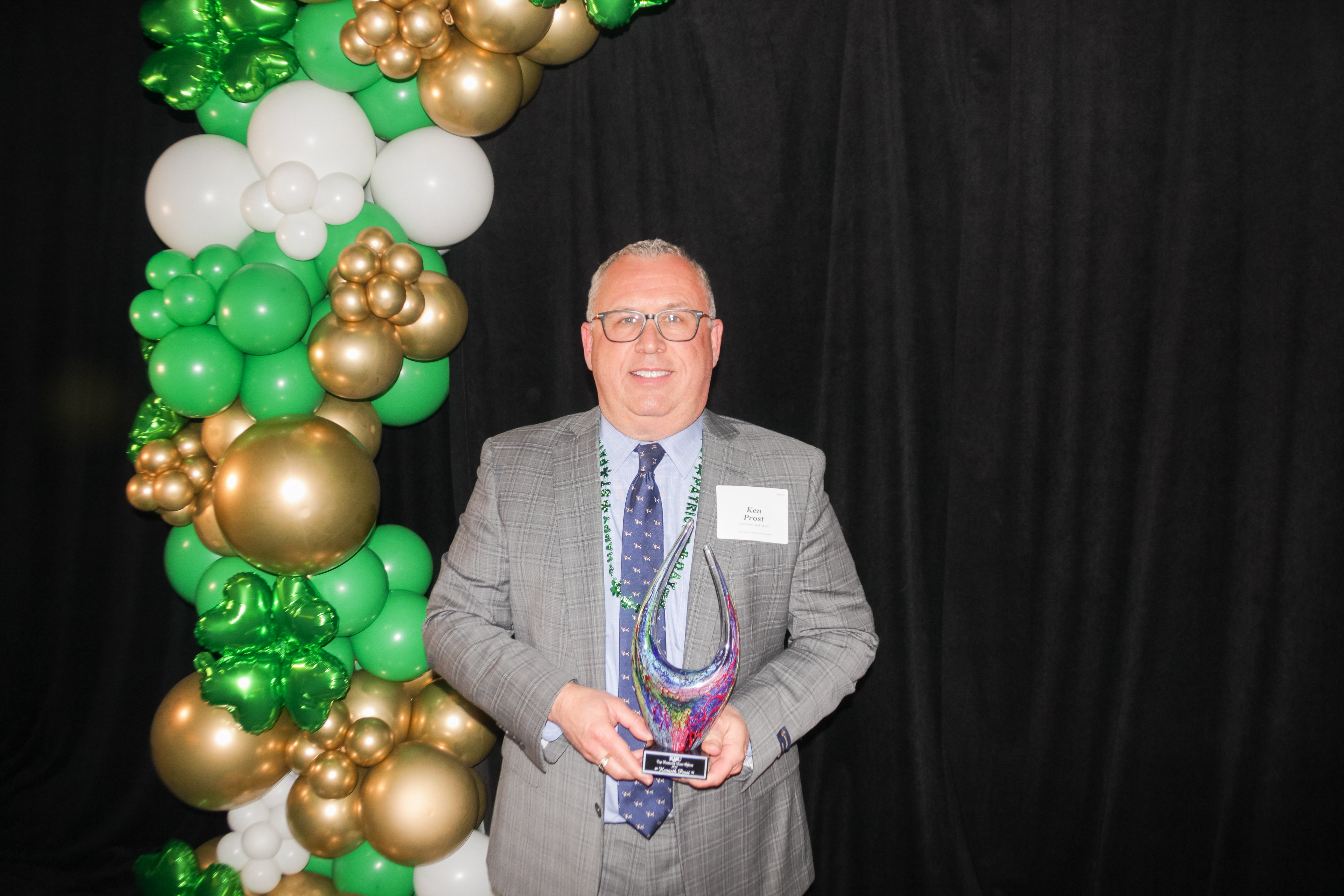 A man in a suit holding an award stands in front of a backdrop with green, gold, and white balloon decorations.