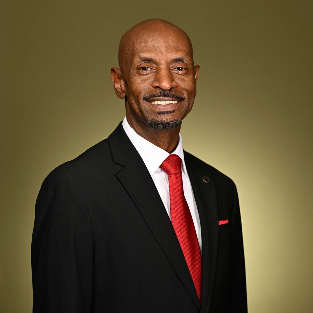 A man in a black suit with a red tie and pocket square stands smiling against a plain olive-green background.