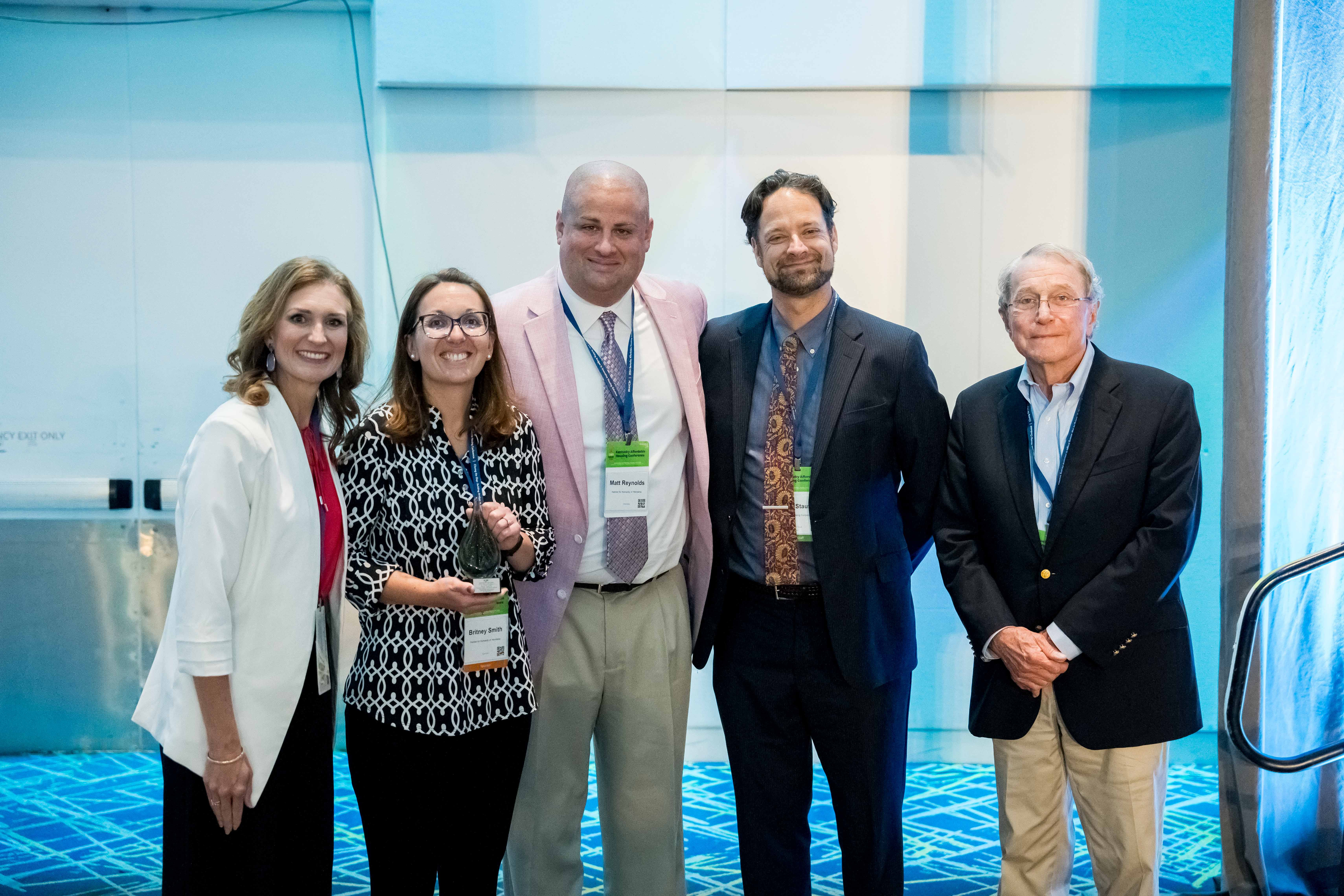 Five people stand indoors at an event, posing for a group photo. They are dressed in business or business-casual attire and smiling at the camera.