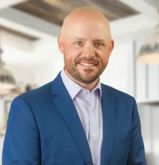 A bald man with a beard, wearing a blue suit jacket and light purple shirt, stands indoors in a modern, well-lit room.