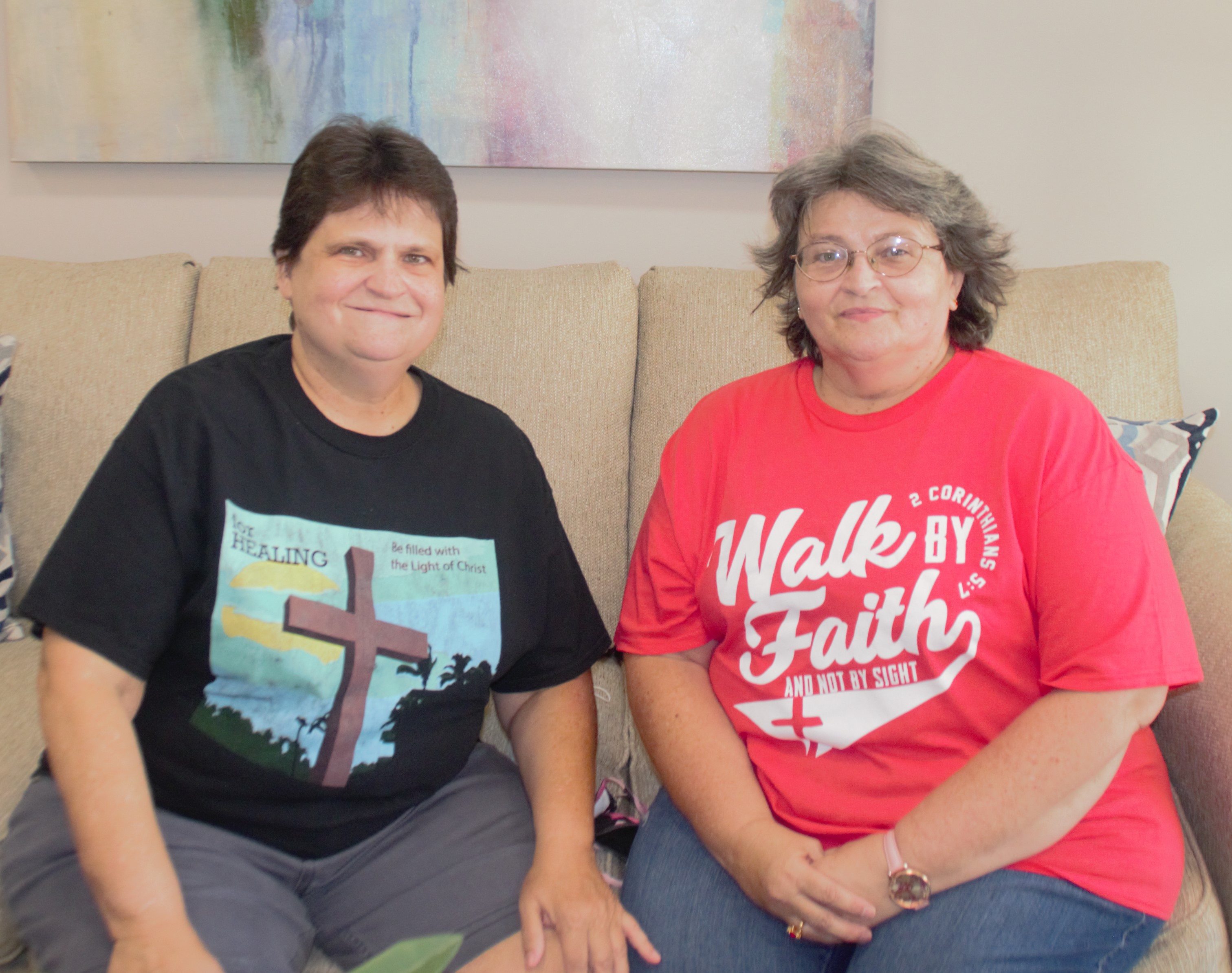 Two women sit on a beige couch, one wearing a black T-shirt with a cross design, the other in a red "Walk by Faith" T-shirt. They both face the camera and smile.