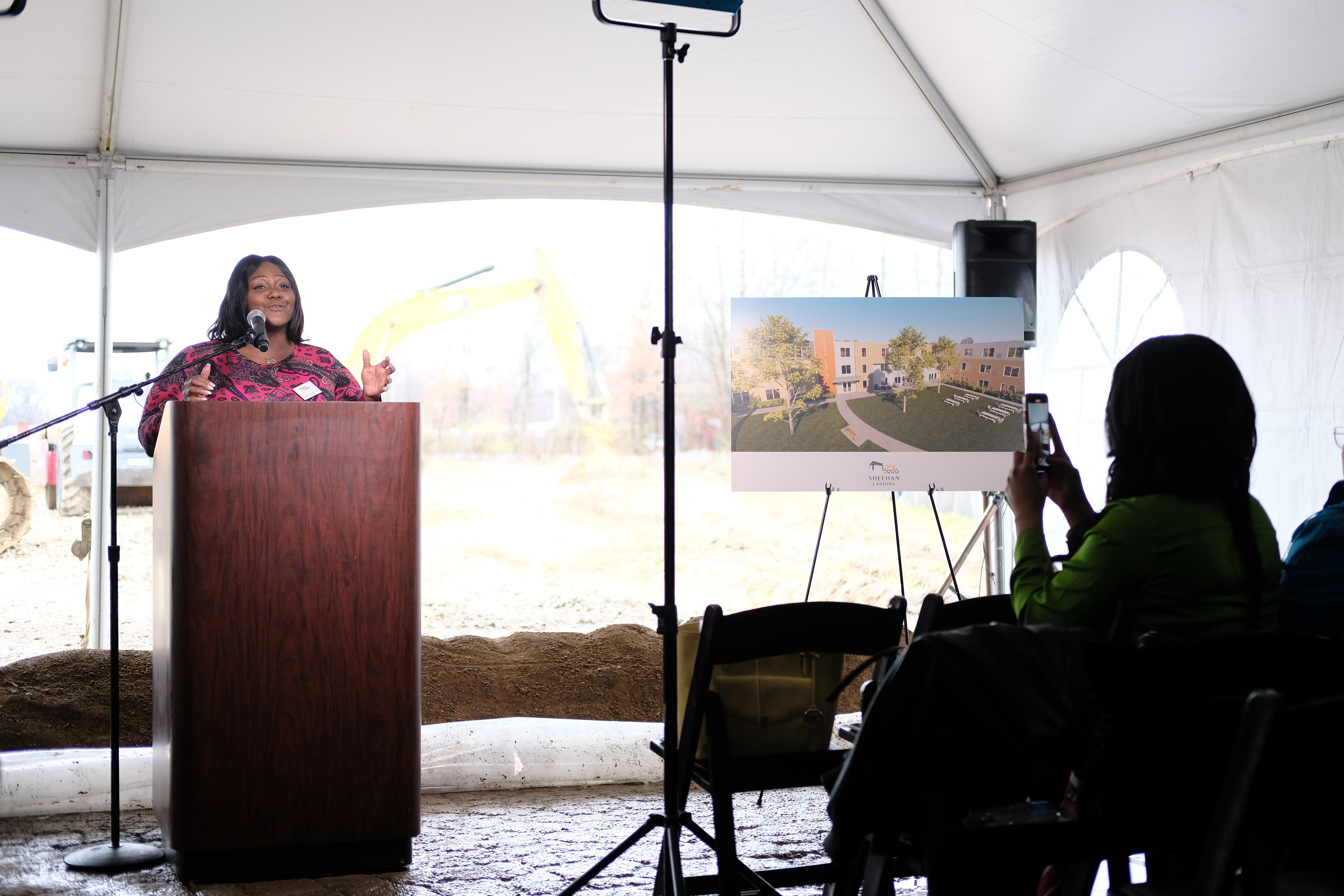 A woman speaks at a podium in a tented event; an audience member takes a photo and a poster with a building rendering is displayed nearby.