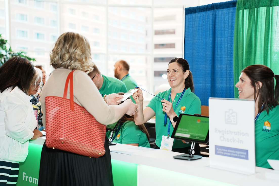 A woman receives a badge from a staff member at a registration check-in desk during an event. Other attendees wait in line beside them.