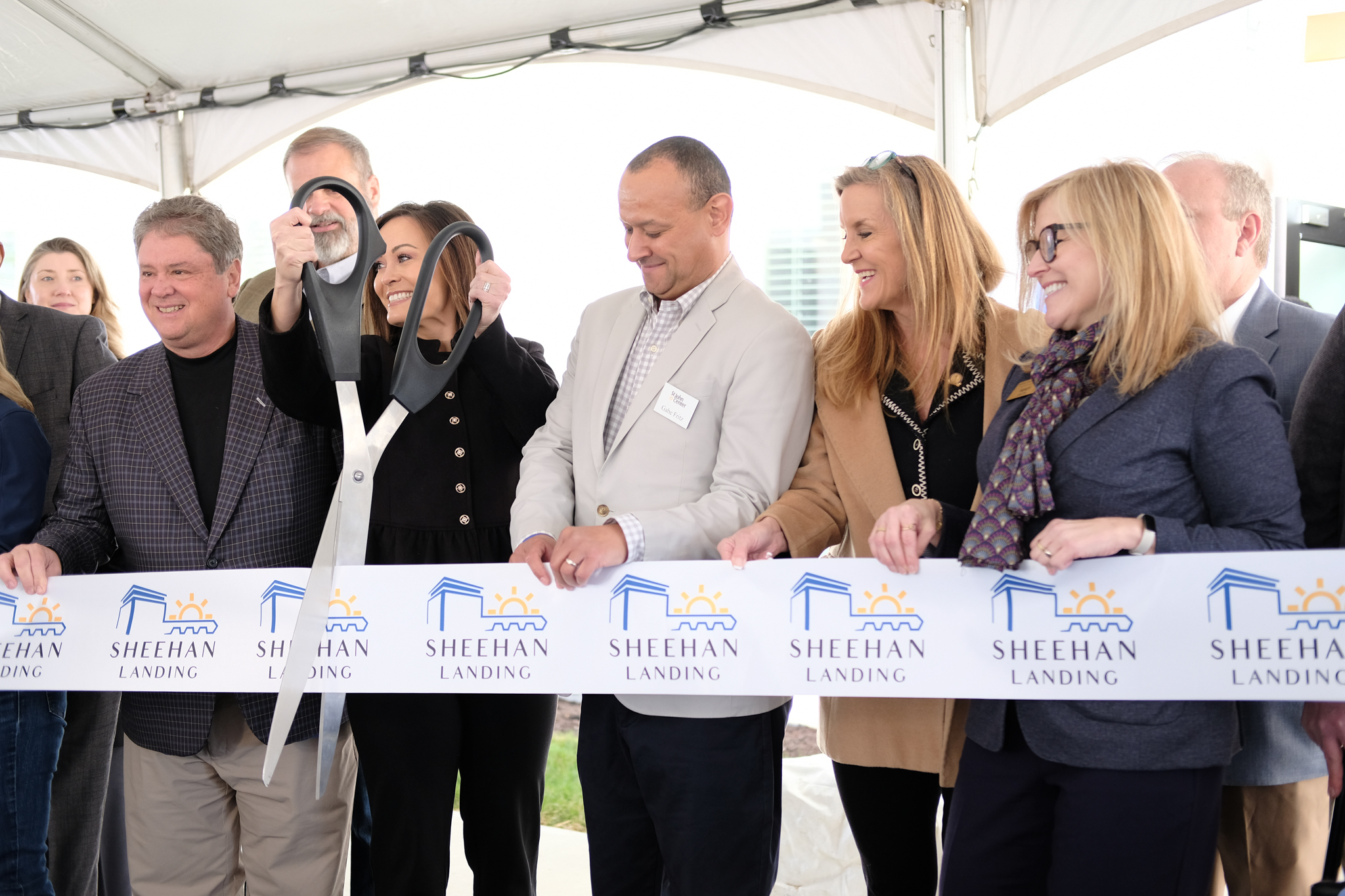 A group of people stand under a tent holding a ribbon labeled "Sheehan Landing" as one person prepares to cut it with oversized scissors.