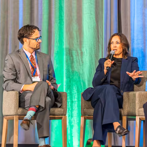 Two people in business attire sit on stage chairs; the woman speaks into a microphone while the man listens attentively. A green and blue curtain serves as the background.