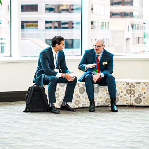 Two men in business suits sit on a patterned bench in an office lobby, engaged in conversation. One has a backpack on the floor next to him. Large windows show city buildings outside.