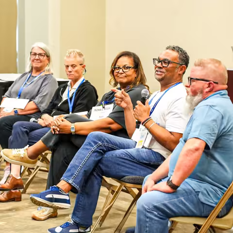 Five people sit on chairs in a panel discussion setting. One person speaks into a microphone while others listen. All wear conference name tags and lanyards.