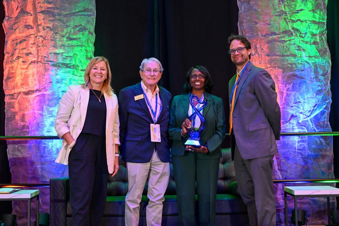Four people pose onstage; one woman in the center holds a blue glass award. The background is lit with multicolored lights.